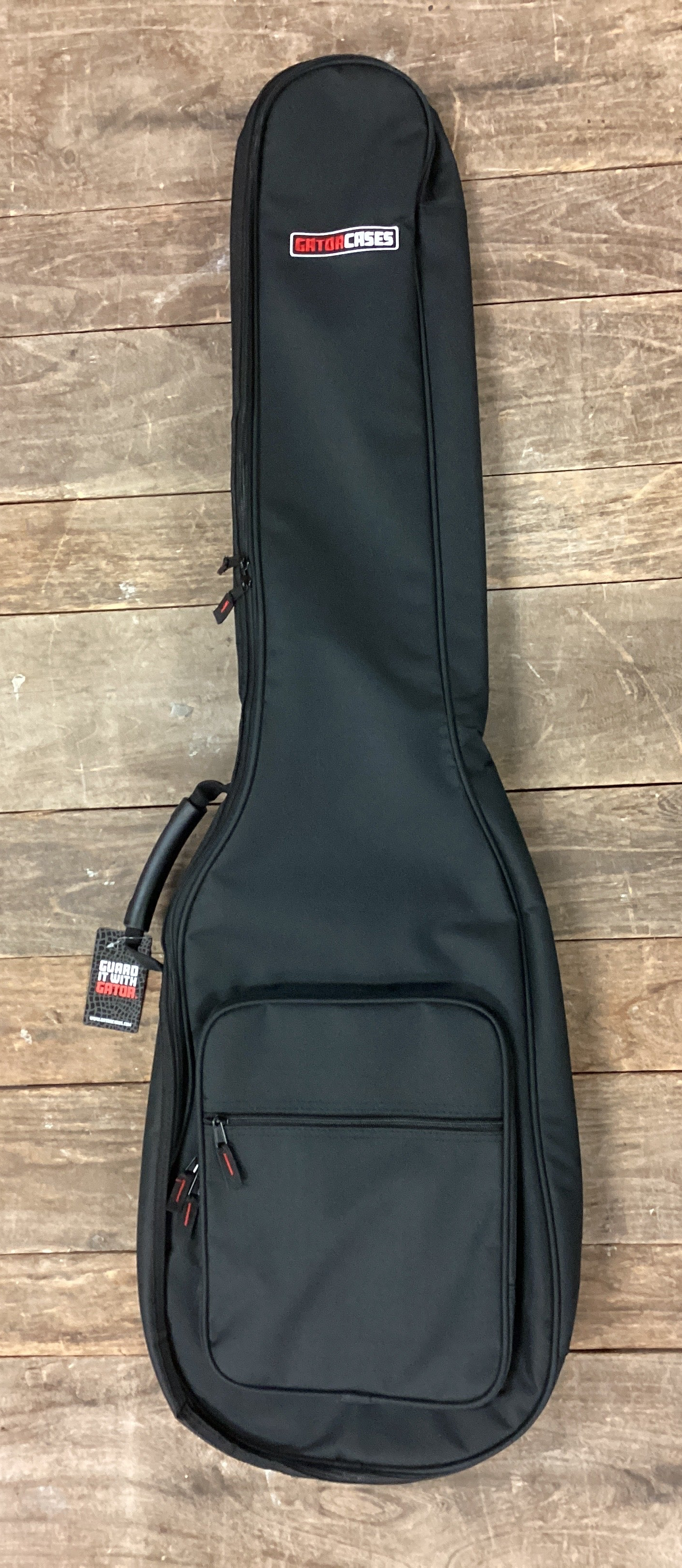 Image of A black guitar or bass gig bag with a front zippered pocket, laid flat on a rustic wooden plank floor. The gig bag features a small red and white logo patch on the side.