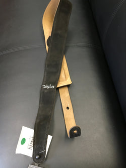 Image of A black leather guitar strap with suede back and Taylor logo embossed on it, placed on a leather surface with a Taylor brand tag attached.