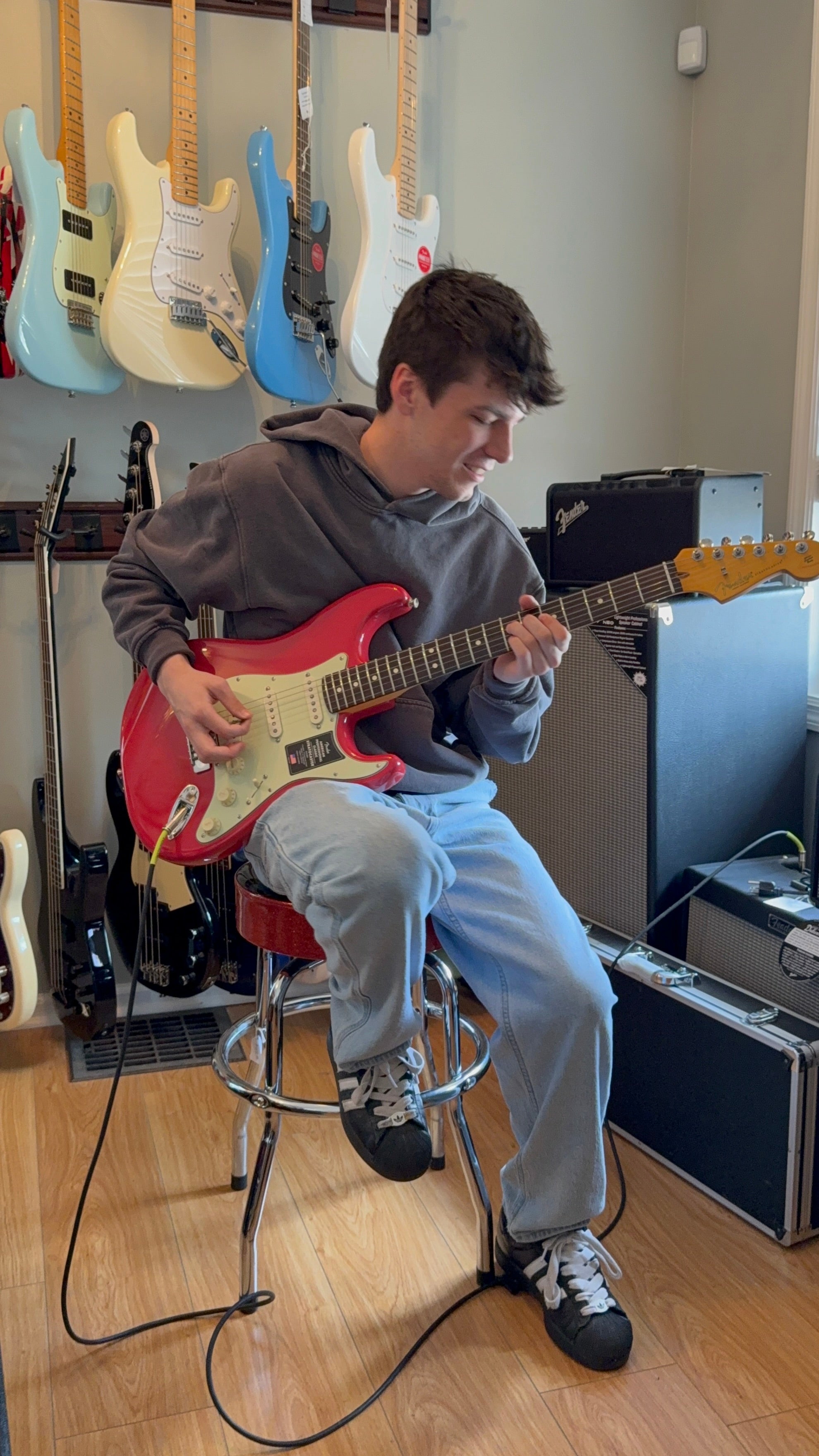 Image of Person playing a red electric guitar in a music store with guitars hanging on the wall.