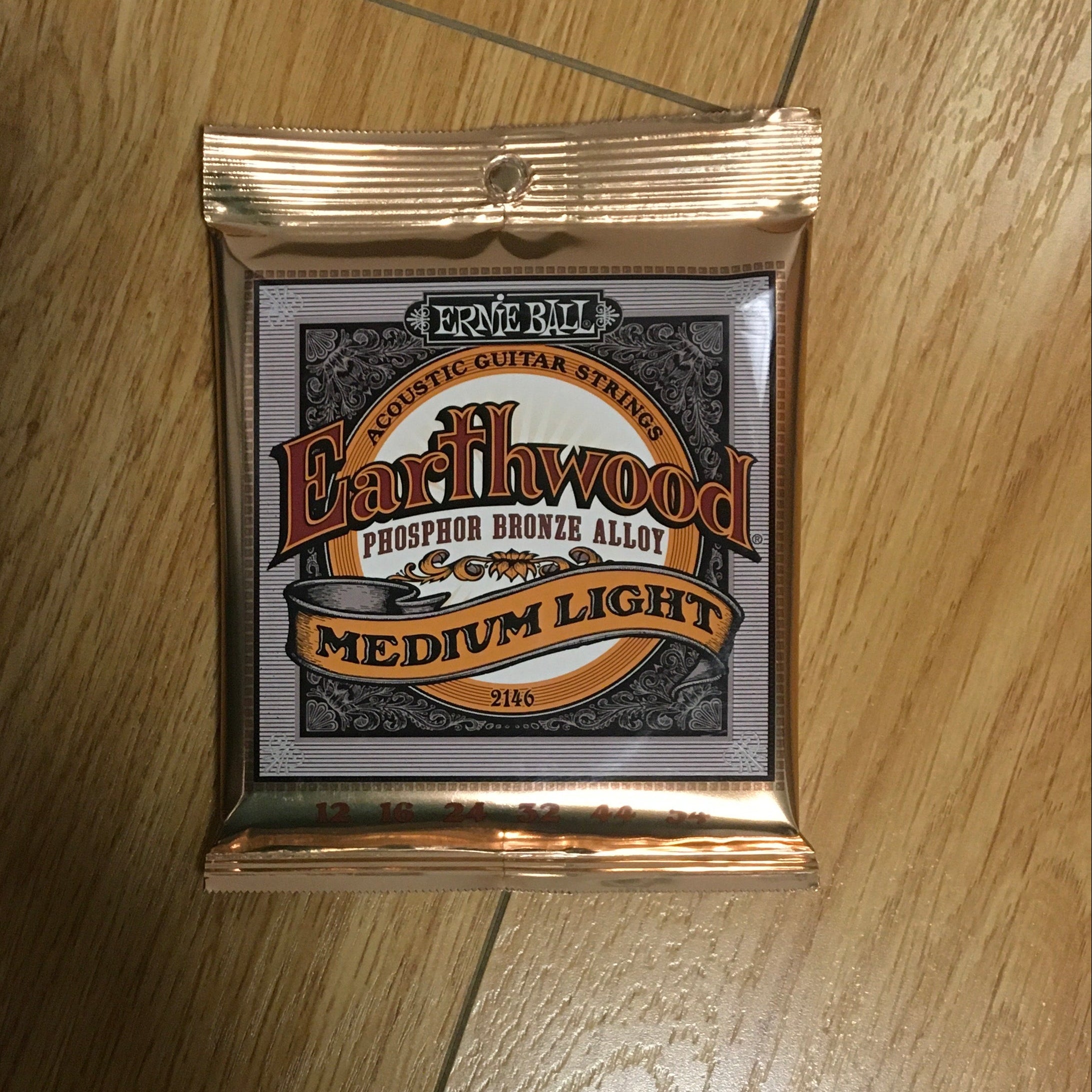 Image of A pack of Ernie Ball Earthwood Phosphor Bronze Medium Light 12-54 Gauge guitar strings placed on a wooden floor.