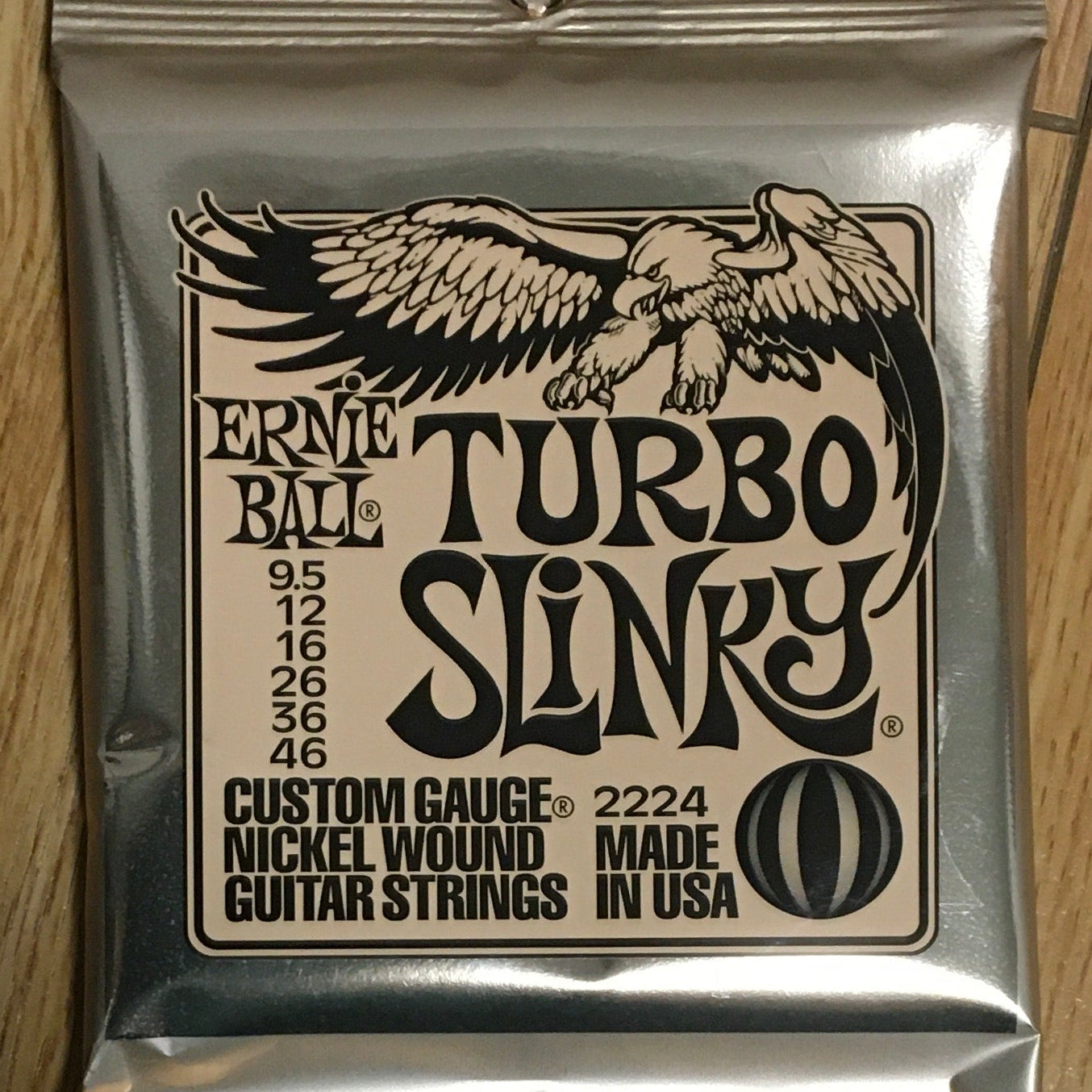 Image of A pack of Ernie Ball Custom Gauge Turbo Slinky guitar strings laid on a wooden floor.