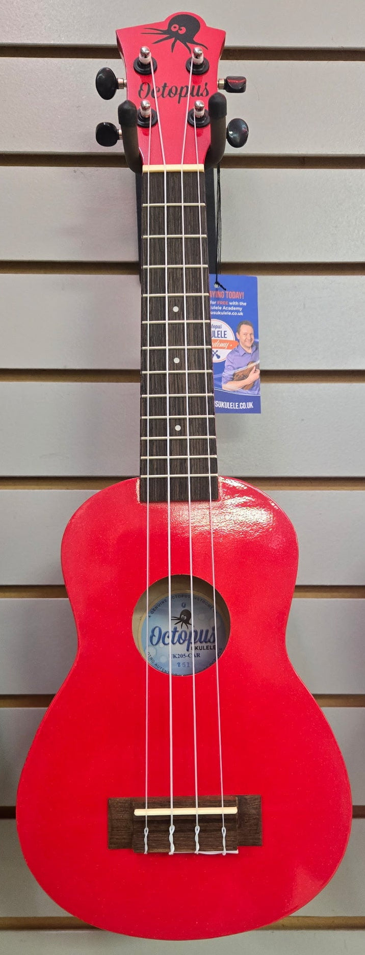 Image of A bright red Octave Ukulele hanging on a slatwall display, with a blue and white price tag attached near the neck.