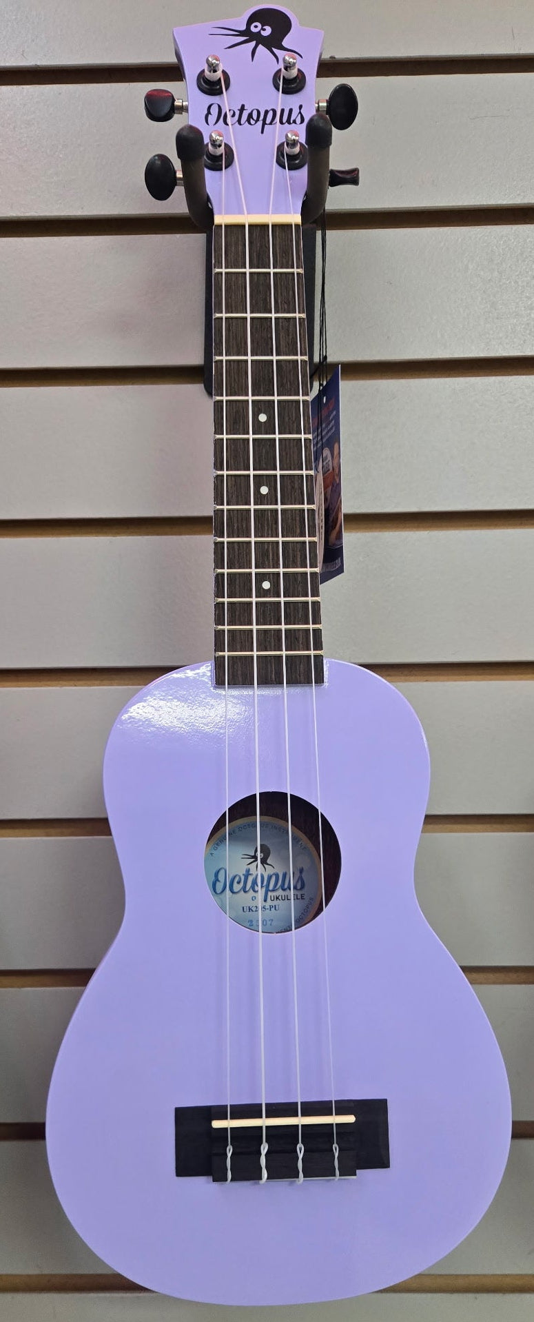 Image of A light purple octetup ukulele hanging on a slatwall display, with other colorful instruments partially visible on the left and right edges.